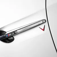 The "M3" emblem and a red V-shaped badge are affixed to the side of a white car, positioned near a sleek side indicator light.