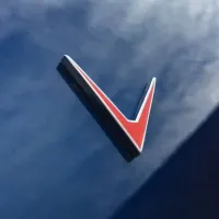 Red, black, and white V-shaped emblem attached to a reflective, shiny blue surface, likely a vehicle exterior. The surrounding area shows blurred reflections and light clouds.
