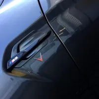 A car door contains a black handle and an emblem displaying "M2" with colored stripes, set against a shiny, dark-colored exterior, reflecting nearby buildings.