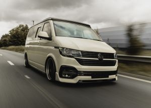 A white van, driving swiftly, is on a multi-lane highway. The background features blurred trees and a guardrail, indicating motion. The weather appears overcast, with a cloudy sky.