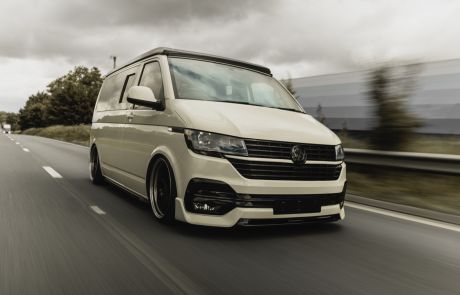 A white van, driving swiftly, is on a multi-lane highway. The background features blurred trees and a guardrail, indicating motion. The weather appears overcast, with a cloudy sky.