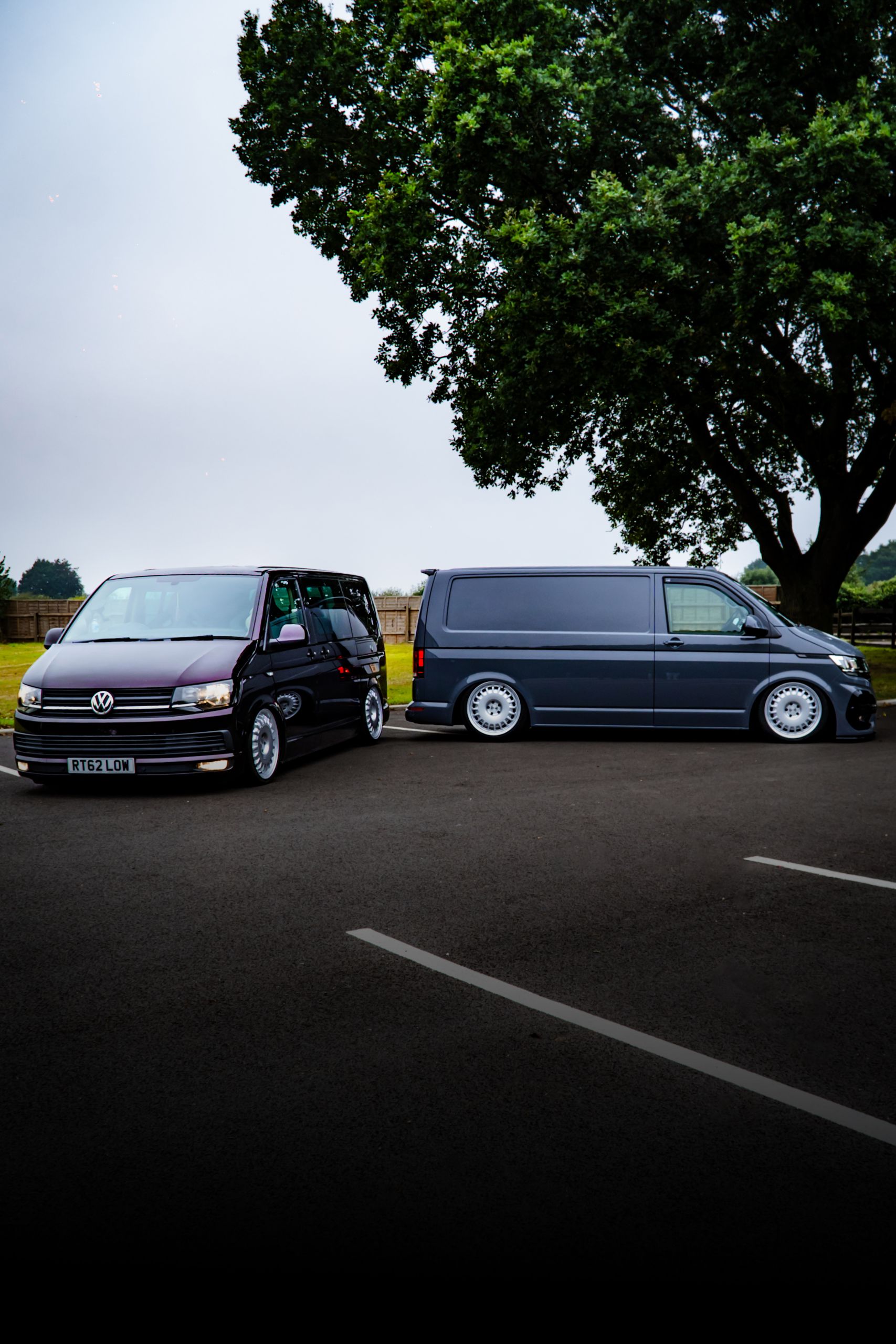 Two Volkswagen vans are parked on an empty asphalt lot beside a large leafy tree. The license plate on one reads, "RT62 LOW."
