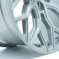 A metallic car wheel rim with intricate geometric patterns rests upright on a white surface, highlighted by soft lighting that accentuates its silver finish.