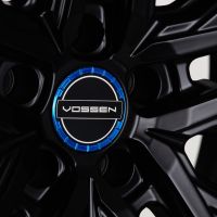 A black car wheel rim features a central logo reading "VOSSEN" surrounded by a striking blue ring, highlighting the branding on the sleek, intricate design.