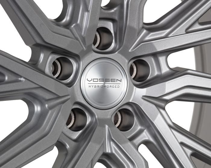 A metallic alloy wheel has intricate, angular spokes radiating from a central hub. Text on the hub reads: "VOSSEN HYBRID FORGED," showcasing the design's precision and branding.