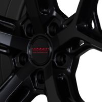 A black alloy wheel features sleek, sharp-edged spokes radiating from a central hub inscribed with "VOSSEN HYBRID FORGED" in red. It is set against a dark background.
