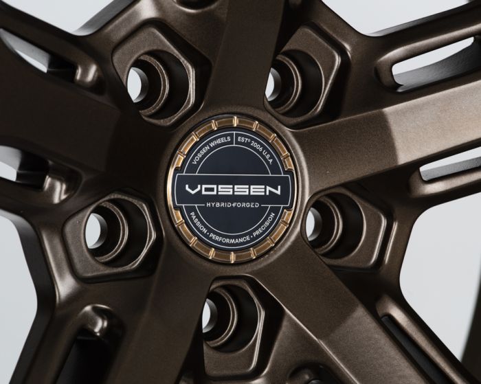 A detailed close-up of a metallic wheel with a central hubcap displaying, "VOSSEN HYBRID FORGED" and text "VOSSEN WHEELS EST. 2006 U.S.A PASSION - PERFORMANCE - PRECISION" in a bright, well-lit setting.