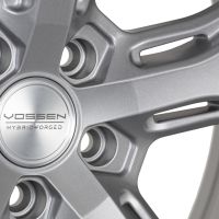 A metallic gray alloy wheel with a central hub cap featuring "VOSSEN HYBRID FORGED" text, surrounded by evenly distributed spokes forming a sleek design.