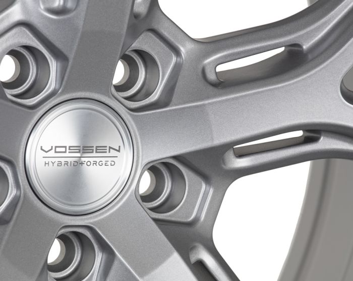 A metallic gray alloy wheel with a central hub cap featuring "VOSSEN HYBRID FORGED" text, surrounded by evenly distributed spokes forming a sleek design.