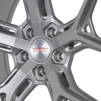 A silver alloy wheel with intricate spokes sits still in a well-lit studio. Text on the center reads: “VOSSEN HYBRID FORGED.”