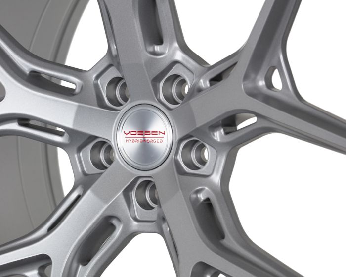 A silver alloy wheel with intricate spokes sits still in a well-lit studio. Text on the center reads: “VOSSEN HYBRID FORGED.”