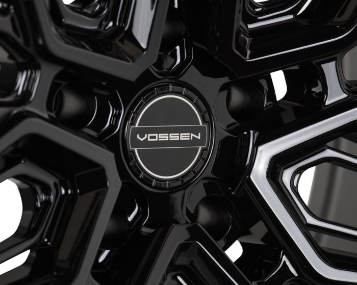 A shiny black alloy wheel features a detailed, intricate pattern with bold geometric lines. The center cap is branded with the word "VOSSEN".