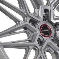 A silver alloy wheel features intricate geometric spokes. The center displays a black circle with the word “VOSSEN” in white, bordered by a red ring, set against a white background.