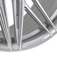 A close-up of a silver alloy wheel with intricate, angled spokes. The text "VOSSEN" is engraved near the rim's edge. The background is a smooth metallic surface.