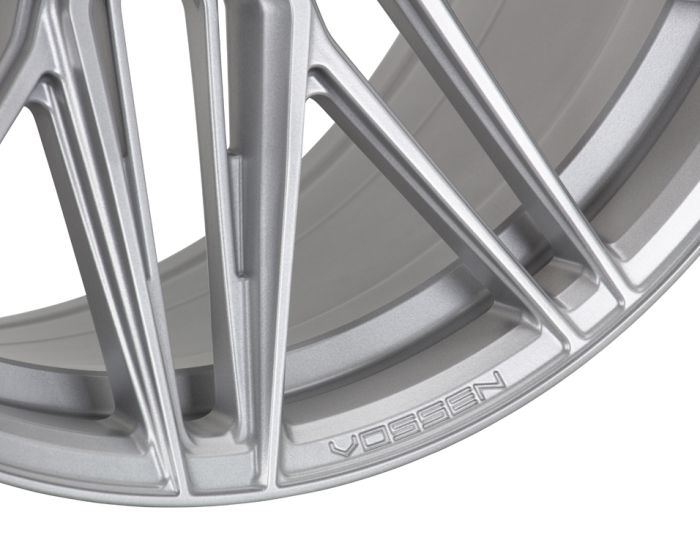 A close-up of a silver alloy wheel with intricate, angled spokes. The text "VOSSEN" is engraved near the rim's edge. The background is a smooth metallic surface.