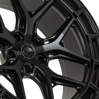 A black car wheel rim features intricate, geometric spokes. The center cap includes text: "VOSSEN FORGED - VOSSEN WHEELS - MIAMI, FL - 6061-T6 FORGED - ENGINEERED ART." The background is plain.
