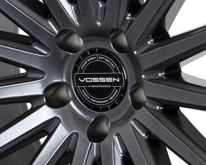 A metallic car wheel with intricate spokes, featuring a central hubcap labeled "Vossen Hybrid Forged." Surrounding text reads: "Vossen Wheels Est. 2006 U.S.A., Passion - Performance - Precision."