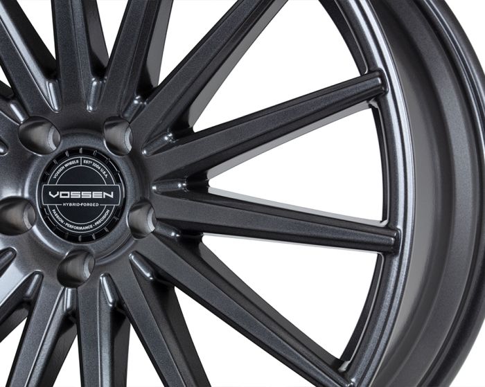 A glossy, dark gunmetal alloy wheel with multi-spoke design features the text "VOSSEN HYBRID FORGED," surrounded by "VOSSEN WHEELS EST 2006 USA" and "PASSION – PERFORMANCE – PRECISION" at the center.