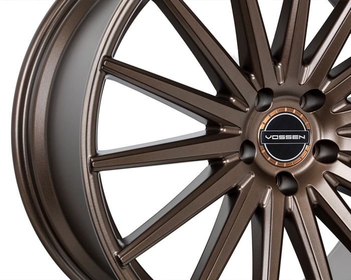 Bronze-colored alloy wheel with intricate spokes radiating from a central hub. The center features a black circle with the text "VOSSEN." The background is white.