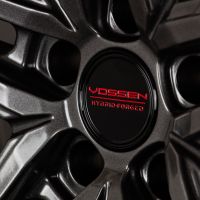 A metallic wheel rim showcases a bold, angular design. The center cap features red text reading: "VOSSEN HYBRID-FORGED." The rim's glossy finish reflects light in a close-up view.
