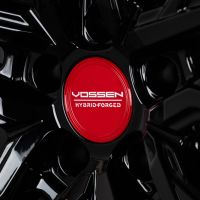 A black wheel rim with a glossy finish features a red center cap labeled "VOSSEN HYBRID FORGED," set against a dark background.