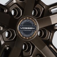 Bronze alloy wheel showcasing intricate design with central logo. Text reads: “VOSSEN WHEELS EST. 2006 U.S.A. VOSSEN HYBRID FORGED PASSION - PERFORMANCE - PRECISION.” Viewed against a neutral background.