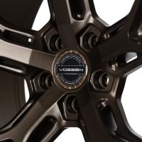 A bronze alloy wheel with a five-spoke design displays the text: "VOSSEN WHEELS EST 2006 U.S.A., VOSSEN, HYBRID-FORGED, PASSION - PERFORMANCE - PRECISION" in a car showroom setting.