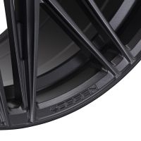 A close-up of a black wheel rim displaying the word "VOSSEN" embossed on the brushed metallic surface, showcasing intricate, angular spokes in a modern, industrial style.