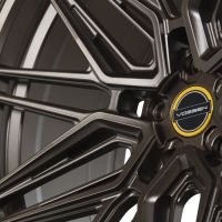An intricate black alloy wheel features multiple spokes, with a central logo displaying "VOSSEN" against a yellow and black circular background; background is neutral, highlighting the wheel's design.