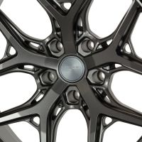 A metallic car wheel rim features intricate, interlacing spokes with a central hubcap. The hubcap displays text: "VOSSEN HYBRID FORGED." The background is a plain, neutral color.