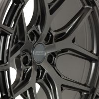 A detailed Vossen hybrid-forged wheel displayed at an angle, showing intricate spoke design, in a studio setting. The wheel has a matte finish with the text "VOSSEN HYBRID FORGED" on the center cap.