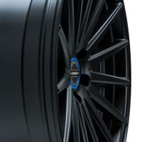 A matte black, multi-spoke car wheel rim is angled, displaying its intricate design. The center features a logo with "VOSSEN FORGED." Bright lighting highlights the finish.