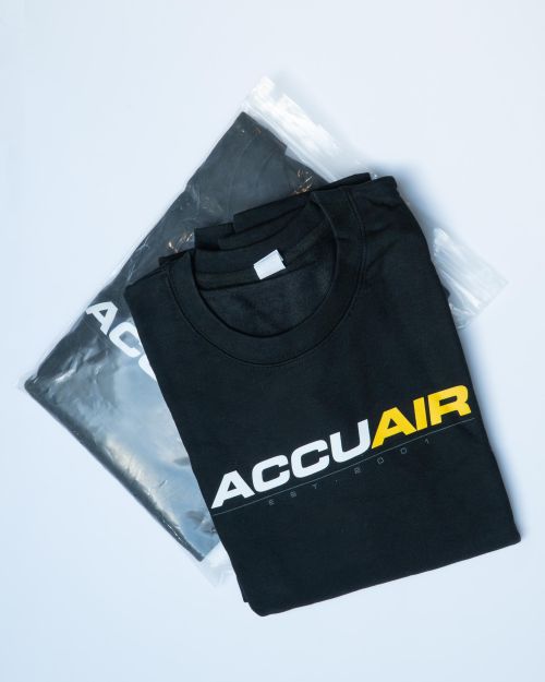 A folded black T-shirt displays the logo "ACCUAIR" in bold letters, with "EST 2001" beneath. Another shirt is partially visible, packaged in a plastic bag, on a white background.