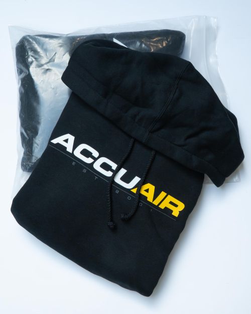 A black hoodie displays "ACCUAIR EST 2001" in bold white and yellow lettering, resting partially out of a clear plastic bag on a white surface.