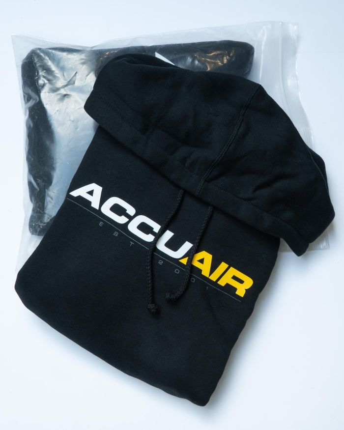 A black hoodie displays "ACCUAIR EST 2001" in bold white and yellow lettering, resting partially out of a clear plastic bag on a white surface.
